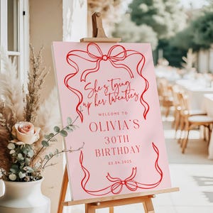 Op de afbeelding: Een roze verjaardagsbord met rode tekst en een rood lintontwerp. De tekst luidt "She's tying up her twenties. Welcome to Olivia's 30th Birthday 03.04.2025".