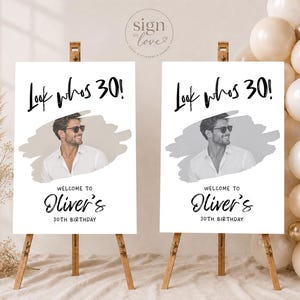 May include: Two white rectangular birthday signs on wooden easels. Each sign features the black text "Look whos 30!", a central photo of a person in a white shirt and sunglasses, and the text "WELCOME TO Oliver's 30TH BIRTHDAY" in black lettering.