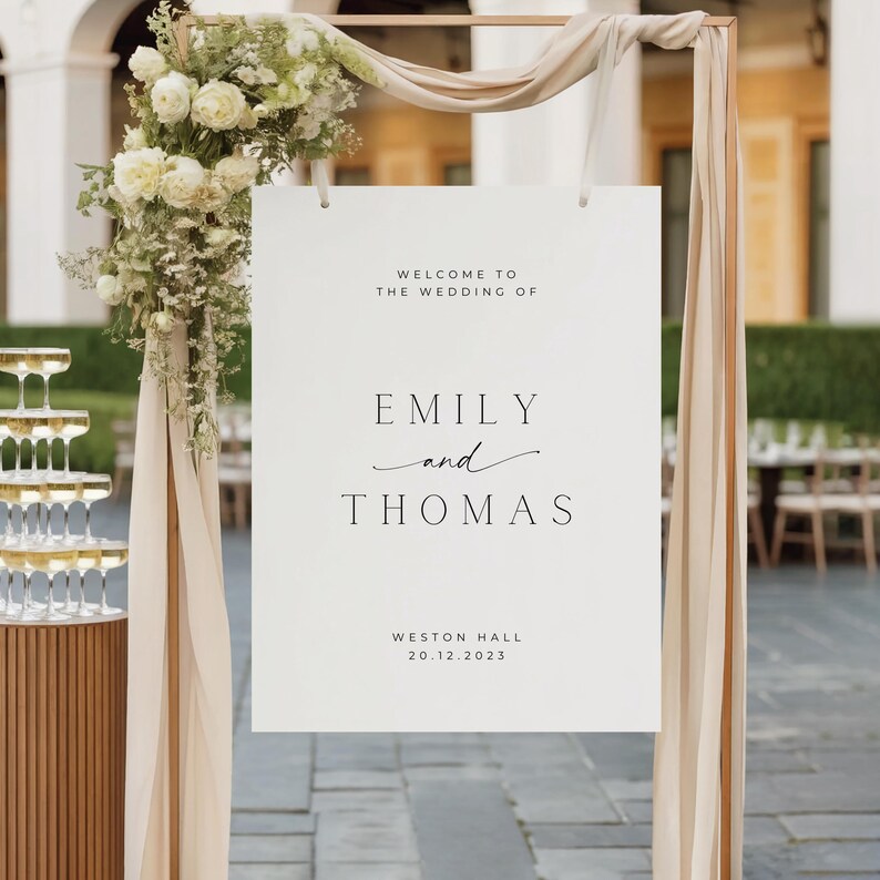 May include: A white wedding welcome sign with the names "Emily and Thomas" in elegant black font. The sign is part of a decorative arch with floral arrangements and draped fabric. The date "20.12.2023" is also displayed.