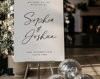 以下が含まれることがあります： ソフィアとジョシュアの結婚式を、2024年12月23日、イスコイドパークにて開催することを告げる、黒い文字で書かれた白い看板。看板は黒いイーゼルに置かれ、その前に3つの銀色のミラーボールが置かれている。