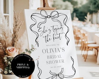 May include: A white sign on a wooden easel with black calligraphy and ribbon designs. The text reads "She's tying the knot," followed by "Welcome to Olivia's Bridal Shower" and the date 03.04.2025. A black circle with text is in the lower left.