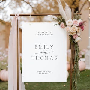May include: A white wedding sign with the text "Welcome to the Wedding of Emily and Thomas" and the date 20.12.2023. The sign is decorated with flowers and greenery, and is part of a wedding decor.