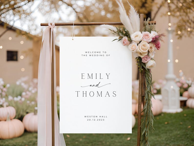 May include: A white wedding sign with the text "Welcome to the Wedding of Emily and Thomas" and the date 20.12.2023. The sign is decorated with flowers and greenery, and is part of a wedding decor.