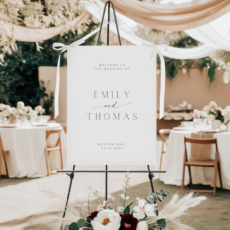 May include: A white wedding welcome sign on a black easel. The sign reads "WELCOME TO THE WEDDING OF EMILY and THOMAS" and "WESTON HALL 20.12.2023". The sign is decorated with white ribbons and sits in front of a floral arrangement.