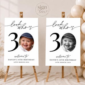 May include: Two identical white rectangular birthday signs on wooden easels. Each sign features the black text 'look who's 30', a large black number '30', a circular photo of a smiling baby in a cap, and the text 'welcome to DAVID'S 30TH BIRTHDAY 16TH MARCH 2024'.