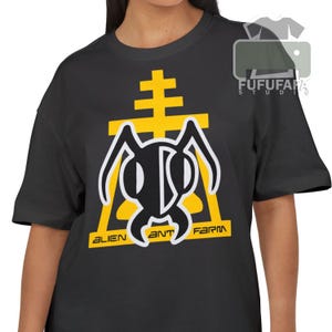 May include: Dark gray t-shirt featuring a yellow and white graphic design. The design includes an ant-like figure with the words "ALIEN ANT FARM" in yellow. The shirt is a crew neck and has short sleeves.
