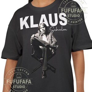 以下が含まれることがあります： 黒いTシャツ。Klaus Schulzeの白黒画像がプリントされています。「KLAUS」という名前が大きな白い文字で画像の上にあり、「Schulze」という名前が筆記体で下に書かれています。このTシャツは綿100％です。