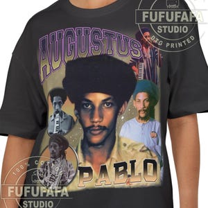 May include: Black t-shirt featuring a vintage-style graphic of Augustus Pablo. The design includes the name "AUGUSTUS" in purple and "PABLO" in orange, along with multiple images of the musician. The shirt has the text "FUFUFAFA STUDIO DTG PRINTED".