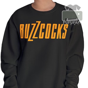May include: Black sweatshirt with the text "BUZZCOCKS" in orange block letters. The sweatshirt has long sleeves and a crew neck. The text is centered on the chest.