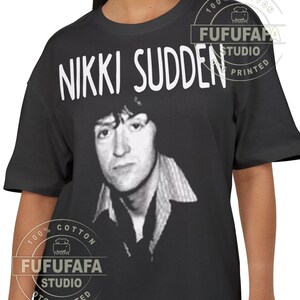 May include: Black t-shirt featuring a black and white portrait of Nikki Sudden with the name "NIKKI SUDDEN" in white block letters above the image. The shirt has a crew neck and short sleeves. The shirt is printed with "FUFUFAFA STUDIO PRINTED" and "100% COTTON" on the front.