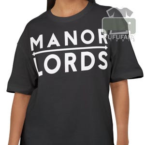 May include: Black t-shirt with the white text "MANOR LORDS" in a bold, sans-serif font. The text is centered on the chest, with a horizontal line separating the two words. The shirt has short sleeves and a crew neck.