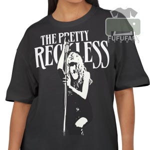 May include: Black t-shirt featuring the band name "The Pretty Reckless" in white, above a black and white illustration of a woman singing into a microphone. The shirt has short sleeves and a crew neck.
