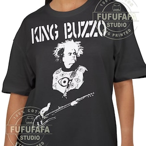 May include: Black t-shirt with a white graphic of a musician and the text "KING BUZZO". The graphic includes a portrait and a guitar. The shirt also has the text "FUFUFAFA STUDIO DTG PRINTED" in a circular design.
