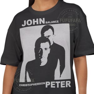 May include: Black t-shirt with a grayscale image of two men. The text "JOHN BALANCE" is above the image, and "CHRISTOPHERSON PETER" is below. The shirt has a crew neck and short sleeves. The shirt is made of 100% cotton.