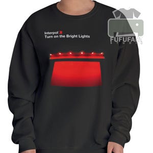 Könnte beinhalten: Schwarzes Sweatshirt mit dem Text "Interpol Turn on the Bright Lights" und einem roten Bühnenmotiv. Das Design zeigt ein rotes Rechteck und eine Reihe kleiner roter Lichter. Das Sweatshirt ist einfarbig schwarz.