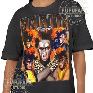 Puede incluir: Camiseta negra con un collage de imágenes de una persona con pelo naranja y maquillaje. La palabra "MARTIN" está impresa en letras grandes y naranjas sobre las imágenes, con "Degville" en cursiva debajo. La camiseta tiene el texto "100% COTTON" y "FUFUFAFA STUDIO DTG PRINTED".