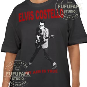 May include: Black t-shirt featuring a black and white image of Elvis Costello playing a guitar. The text "ELVIS COSTELLO" is printed in red across the top, with "AIM IS TRUE" in red below the image. The shirt is made of 100% cotton.