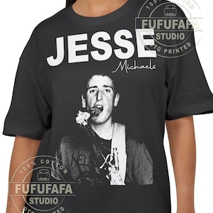 Puede incluir: Camiseta negra con una imagen en blanco y negro de una persona cantando en un micrófono. El nombre "JESSE Michaels" está impreso en blanco encima de la imagen. La camiseta es 100% algodón y tiene el logo "FUFUFAFA STUDIO DTG PRINTED".