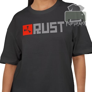 Pode incluir: Camiseta cinza escuro com a palavra "RUST" em letras cinzas. A letra "R" está em um quadrado vermelho com um design estilizado. A camiseta tem mangas curtas e gola redonda.