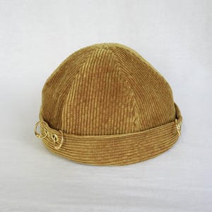 Könnte beinhalten: Ein brauner Cordhut mit abgerundeter Krone und flachem Rand. Der Hut hat zwei goldene Metallringe am Rand.