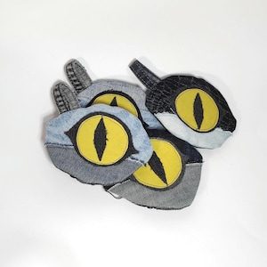 Könnte beinhalten: Vier handgefertigte Katzenaugenspielzeuge aus Jeansstoff. Jedes Spielzeug hat ein gelbes Auge mit einer schwarzen Pupille.