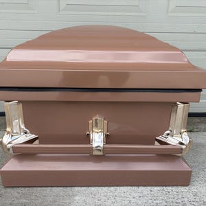 May include: A bronze-colored casket with a glossy finish. The lid has a curved design, and the body features gold-colored handles and accents. The casket is designed for burial and has a rectangular shape.