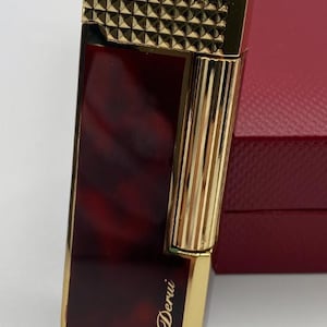 May include: A gold-coloured lighter with a geometric pattern on the top and vertical lines on the side. The body is a reddish-brown colour with a marbled effect. The word "Derui" is written in gold script on the front.