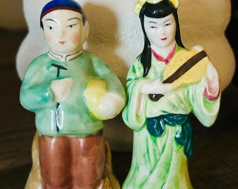 Könnte beinhalten: Ein Paar antike Porzellanfiguren, die einen Mann und eine Frau in traditioneller chinesischer Kleidung darstellen. Der Mann hält einen gelben Ball und einen Stab, während die Frau einen Fächer hält. Die Figuren sind in leuchtenden Farben bemalt, darunter Grün, Rot, Gelb und Blau.