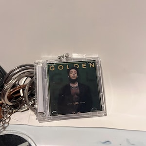 Könnte beinhalten: Ein durchsichtiger Kunststoff-Schlüsselanhänger mit einem CD-Case mit dem Wort "GOLDEN" in goldfarbenen Buchstaben. Das CD-Cover zeigt ein Foto einer Person. Der Schlüsselanhänger ist an einem silbernen Schlüsselring befestigt.