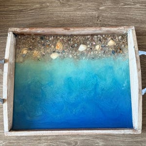 May include: A rectangular wooden serving tray with a beach-themed design. The tray's interior features a blue and turquoise resin design with seashells and small pebbles. The tray has metal handles and a weathered wood frame.