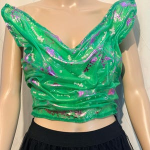 Può includere: Un top corto verde brillante con scollatura drappeggiata e dettagli sulle spalle. Il top presenta accenti viola iridescenti e piccoli abbellimenti bianchi. Il fondo è abbinato a una gonna nera.
