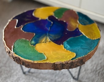 May include: A round wooden side table with a colorful epoxy resin top. The top features a mosaic design with shades of blue, green, yellow, and brown, outlined in gold. The table is supported by three black metal hairpin legs.