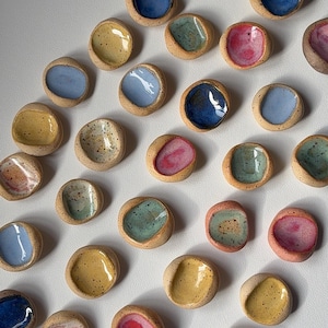 Puede incluir: Una colección de pequeños cuencos de cerámica hechos a mano en varios colores. Los cuencos tienen forma redondeada con un interior poco profundo y coloreado. Los colores incluyen azul, amarillo, rosa y verde. Los cuencos están dispuestos sobre una superficie blanca.