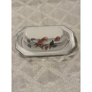 Puede incluir: Una mantequillera rectangular de cerámica con tapa, que presenta un color base blanco roto y un delicado patrón floral en tonos rosa y verde. El plato tiene un borde de color azul oscuro a lo largo de los bordes tanto de la base como de la tapa.