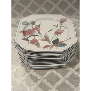 May include: A stack of octagonal white ceramic plates featuring a floral pattern with muted pink flowers and dark green leaves. Each plate has a thin dark gray rim along the edge, creating a uniform look for this set of dinnerware.