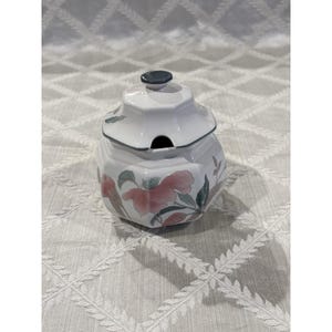 Puede incluir: Un azucarero de cerámica blanca con tapa a juego, que presenta una forma octogonal de múltiples facetas. La superficie está decorada con un delicado patrón floral de flores rosas y hojas verdes, acentuado por un borde gris oscuro en la tapa.