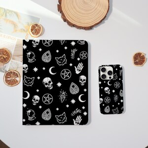 May include: Black phone case and notebook featuring a white pattern of skulls, cats, stars, and occult symbols. The phone case is next to the notebook, both on a white surface.