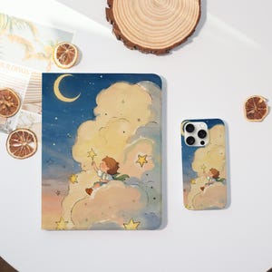 May include: A notebook and phone case featuring a whimsical illustration of a child reaching for a star in a night sky with clouds. The color palette includes blues, yellows, and oranges. The notebook and phone case are on a white surface.