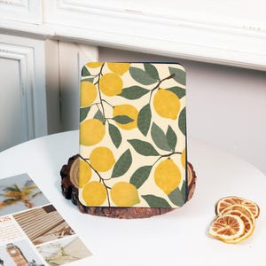 Może przedstawiać: Prostokątne etui z wzorem gałęzi cytryny, przedstawiające żółte cytryny i zielone liście na kremowym tle. Etui jest umieszczone na drewnianym plastrze, z suszonymi plasterkami cytryny w pobliżu. Projekt przywodzi na myśl świeży, letni nastrój.
