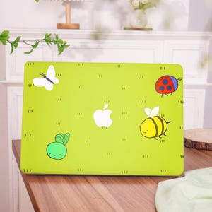 Könnte beinhalten: Eine hellgrüne Laptop-Hülle mit einem weißen Apple-Logo und Cartoon-Illustrationen eines Schmetterlings, Marienkäfers, einer Biene und einer Schnecke. Die Hülle hat ein grasartiges Muster mit kleinen vertikalen Linien.