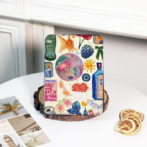 May include: A decorative case featuring a collage of colorful illustrations. The design includes a disco ball, goldfish, berries, a bottle of gin, and the text "Can we get a bread basket?" on a light yellow background.