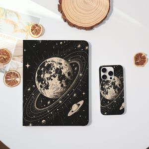 May include: A black and white celestial-themed phone case and tablet case. The cases feature a detailed illustration of a planet with rings, surrounded by stars, on a black background. The cases are displayed on a white surface.