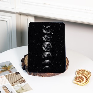 May include: A black tablet case featuring a celestial design of the moon phases against a starry background. The case is rectangular and rests on a wooden slice, with a white table and decorative elements in the background.