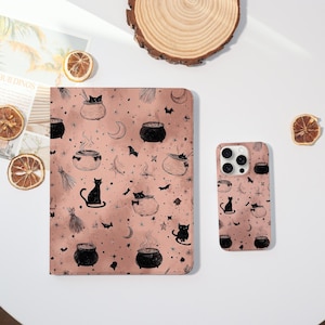 May include: A pink tablet case and phone case featuring a repeating pattern of black cauldrons, cats, bats, moons, and stars. The cases are on a white surface with dried orange slices and a wooden slice.