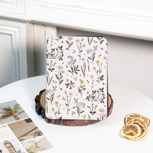 Könnte beinhalten: Ein rechteckiger Notizbuchumschlag mit floralem Muster. Der Umschlag hat einen weißen Hintergrund mit einem sich wiederholenden Muster aus gelben, blauen und violetten Wildblumen. Das Notizbuch liegt auf einer Holzscheibe.