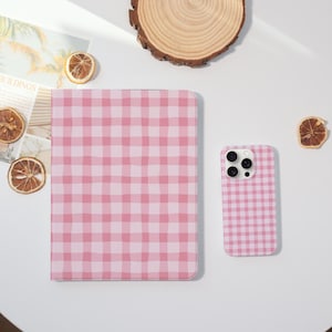 May include: A pink and white gingham pattern adorns a tablet case and a smartphone case. The cases are displayed on a white surface, alongside dried orange slices and a wooden coaster. The overall aesthetic is bright and cheerful.