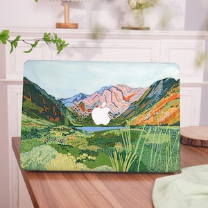May include: A laptop skin featuring a painted design of a mountain range with a lake in the valley. The mountains are painted in shades of pink, blue, green, and orange. The lake is a light blue colour. The design is surrounded by green grass and trees.