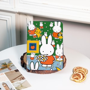 Op de afbeelding: Een groene, blauwe en oranje hoes met een herhalend patroon van witte konijnen die bloemen vasthouden, een gele hond en een oranje appel. De hoes is ontworpen voor een e-reader of tablet.