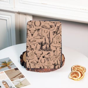 May include: A tan-colored Kindle cover with a black and white pattern of cowboy boots, cacti, horseshoes, and skulls.