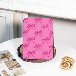 May include: A pink tablet case featuring a repeating pattern of pink bows. The case is displayed on a wooden base, set against a white background. The bows are a lighter shade of pink than the case's background.